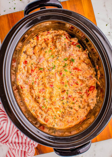 A crockpot filled with a chunky, creamy dip featuring visible pieces of tomato and herbs, placed on a wooden board with a red and white striped cloth nearby.