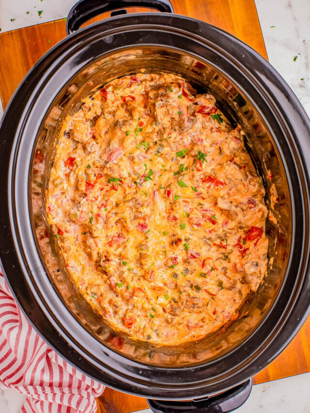 A crockpot filled with a chunky, creamy dip featuring visible pieces of tomato and herbs, placed on a wooden board with a red and white striped cloth nearby.