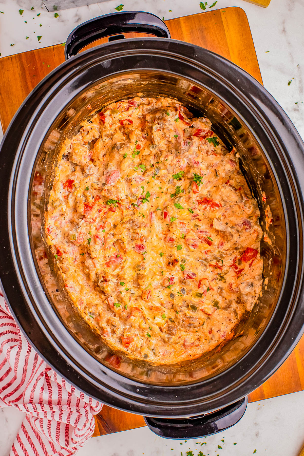 A crockpot filled with a chunky, creamy dip featuring visible pieces of tomato and herbs, placed on a wooden board with a red and white striped cloth nearby.