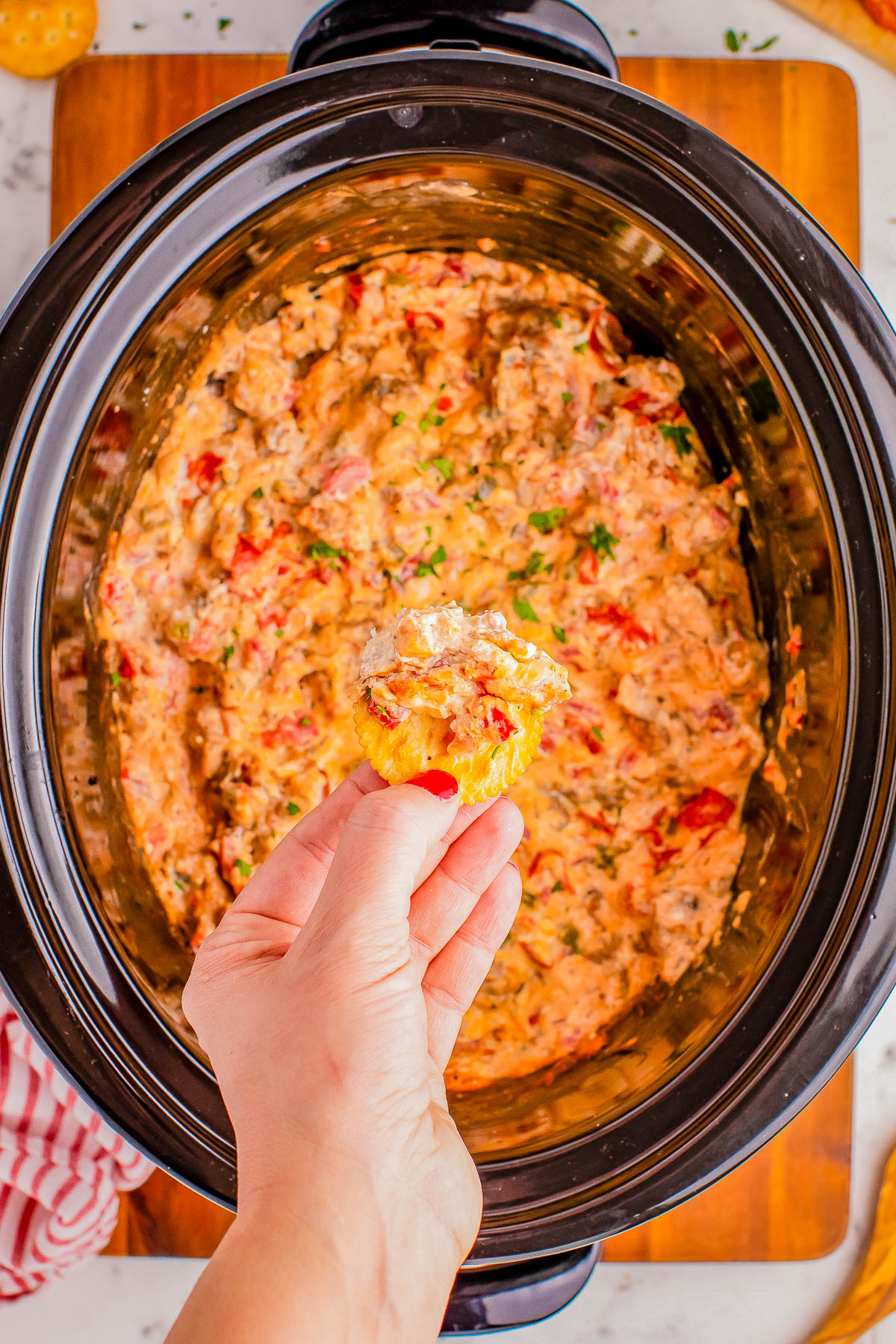 A person holds a chip with a dip in front of a black slow cooker filled with a creamy, chunky dip that includes red and green ingredients.