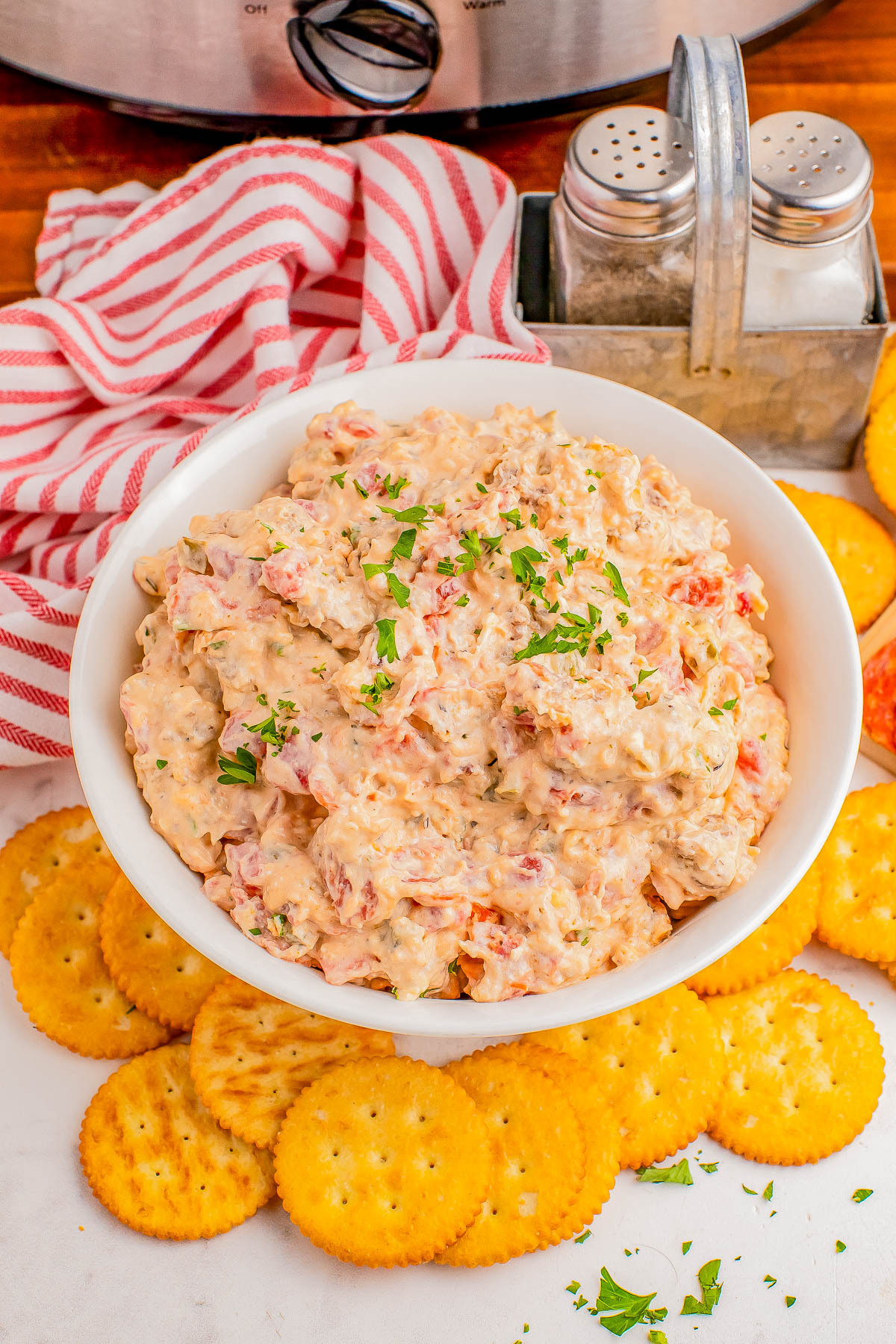 A white bowl filled with a creamy dip topped with chopped herbs, surrounded by round crackers on a white surface. A striped red and white cloth and a salt and pepper set are in the background.