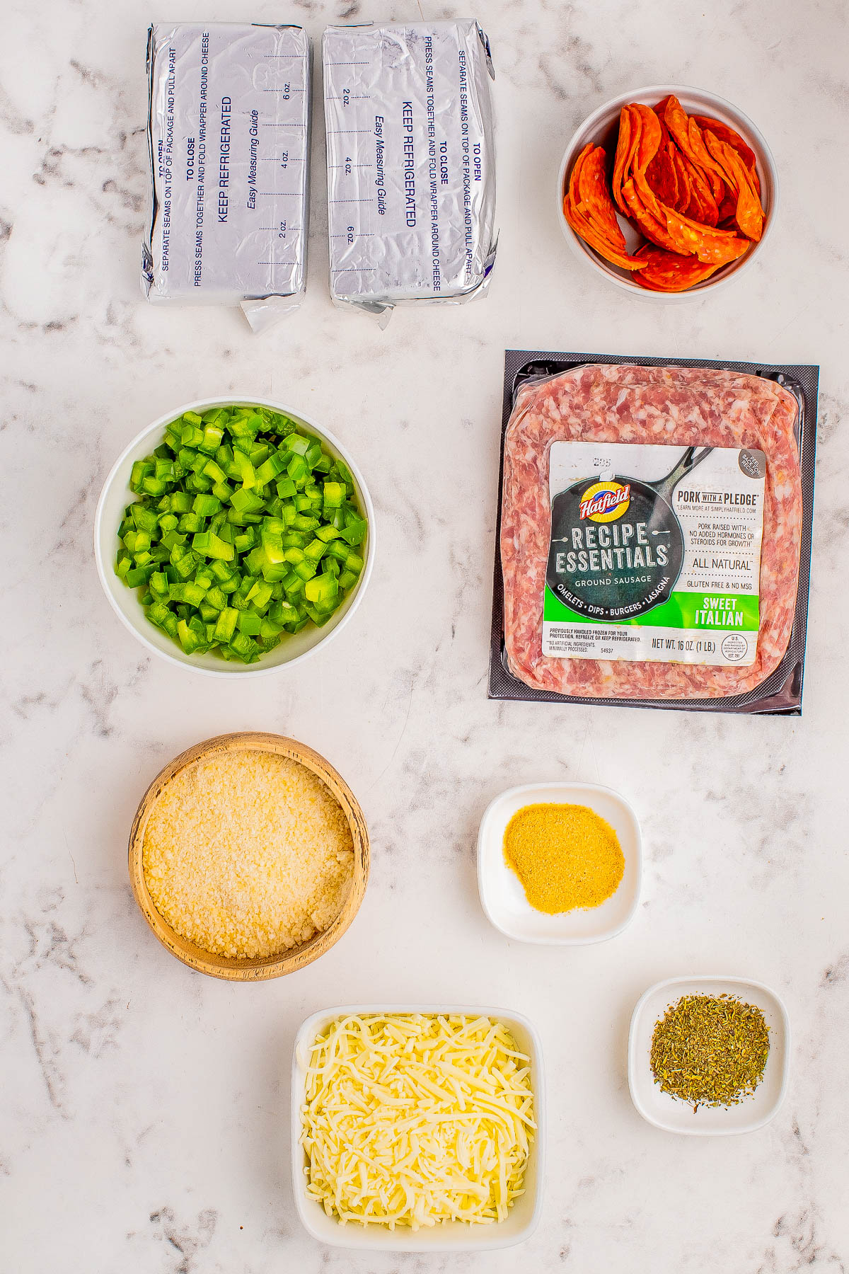 Ingredients on a marble surface include two packages of cream cheese, pepperoni slices, chopped green bell peppers, ground pork, breadcrumbs, shredded cheese, cornmeal, and dried herbs.