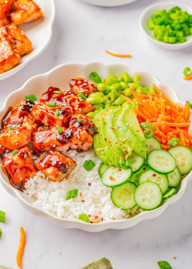 A bowl of rice topped with glazed salmon, sliced avocado, shredded carrots, sliced cucumber, edamame, and garnished with chopped green onions and sesame seeds.