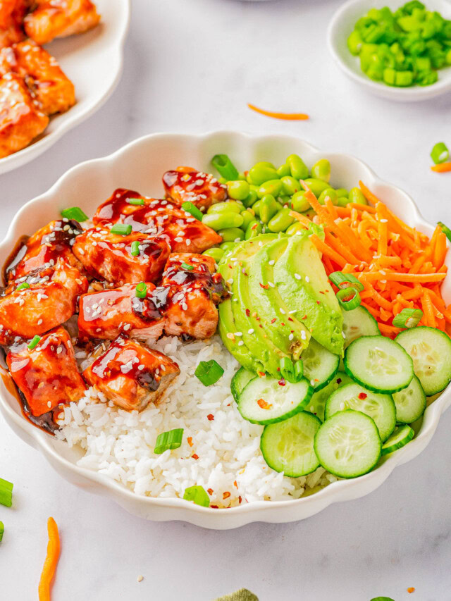 A bowl of rice topped with glazed salmon, sliced avocado, shredded carrots, sliced cucumber, edamame, and garnished with chopped green onions and sesame seeds.