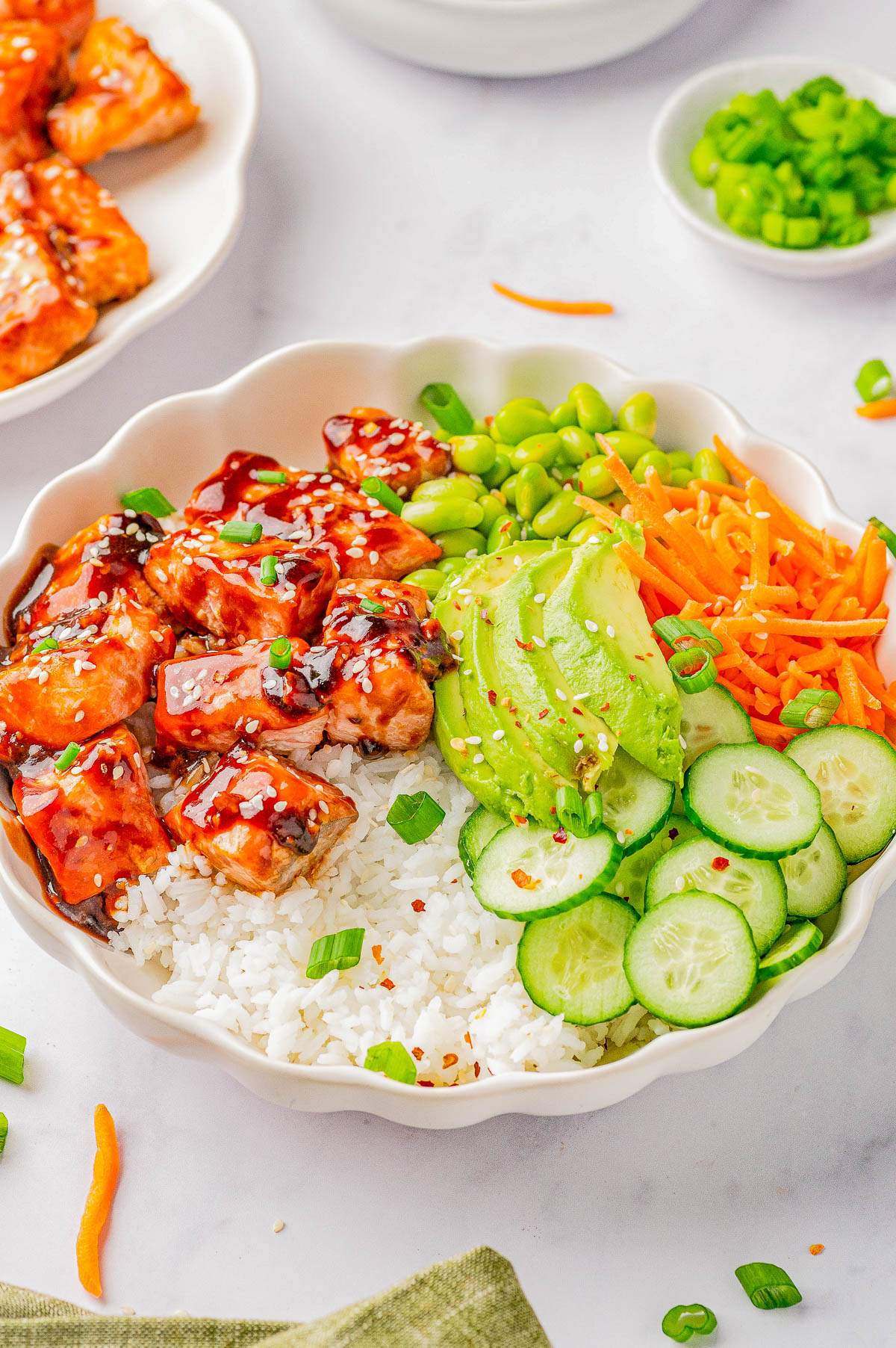 A bowl of rice topped with glazed salmon, sliced avocado, shredded carrots, sliced cucumber, edamame, and garnished with chopped green onions and sesame seeds.