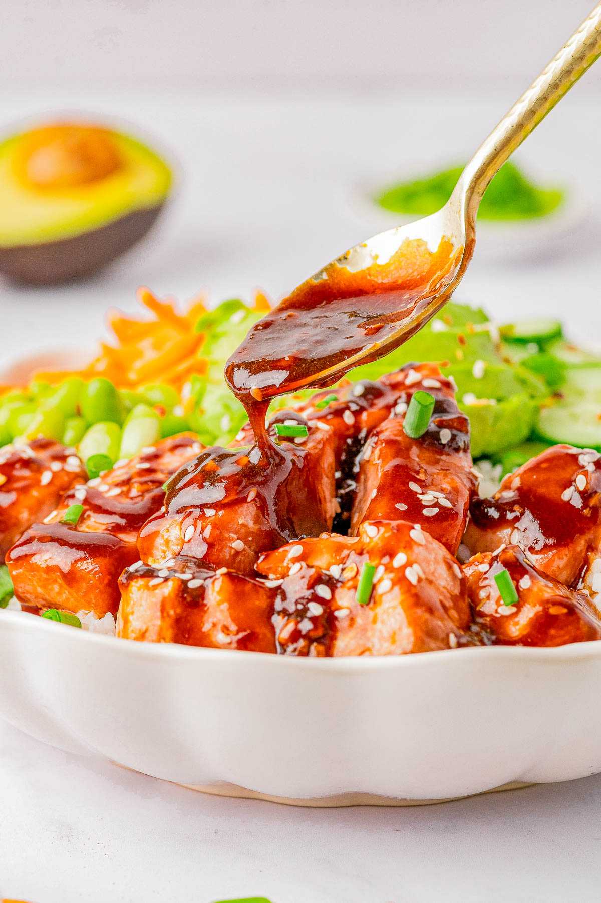 A spoon drizzling teriyaki sauce over a bowl of grilled chicken pieces, garnished with sesame seeds and chives, with sliced vegetables and half an avocado in the background.