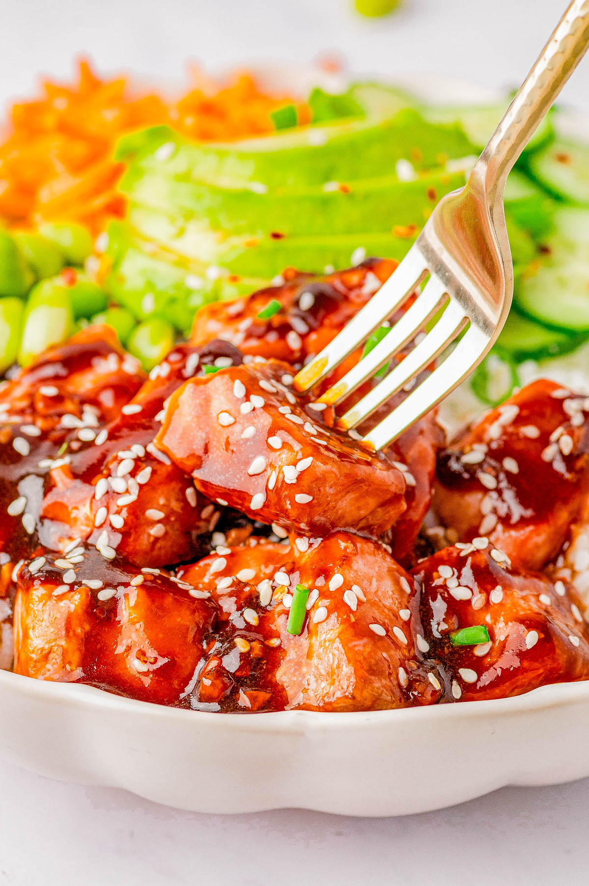 A bowl of glazed teriyaki salmon topped with sesame seeds, paired with sliced avocado, cucumber, and diced vegetables. A fork is lifting a piece of chicken.