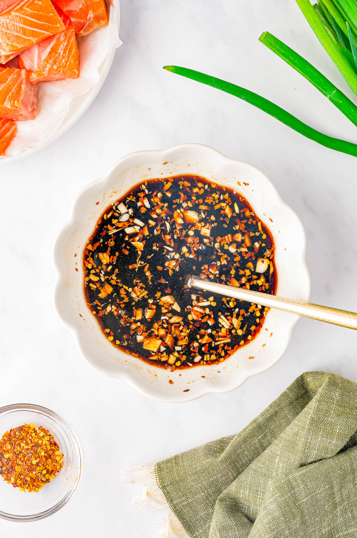 A bowl filled with a dark soy sauce mixture with chopped garlic and spices, next to a plate of raw salmon, a green onion, and a bowl of red pepper flakes on a white surface.