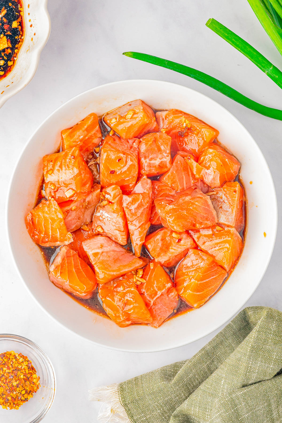 A bowl of raw diced salmon marinated in a reddish sauce, placed on a light-colored surface, with green onions, a green cloth napkin, and a small dish of red pepper flakes nearby.