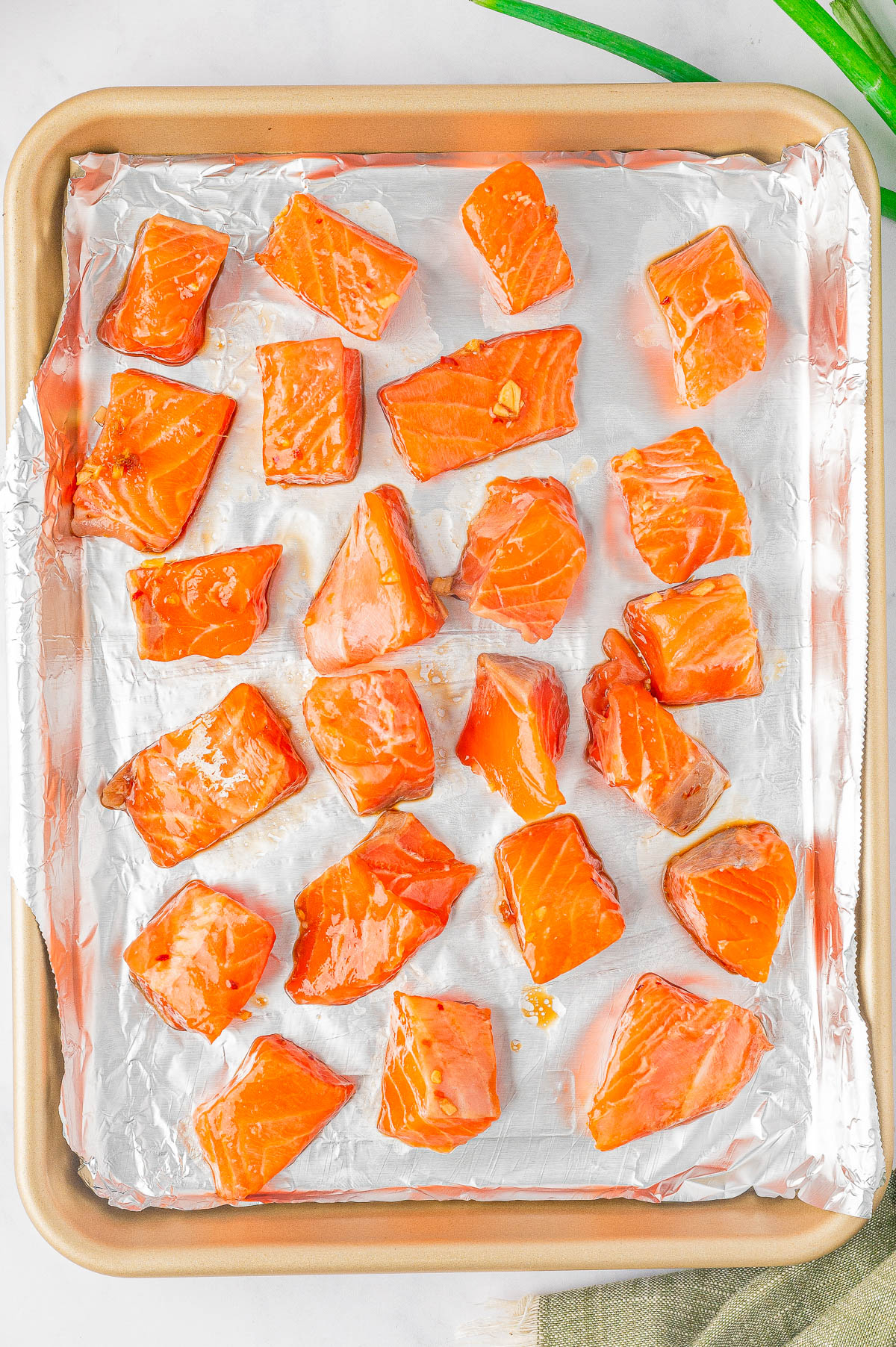 A baking sheet lined with aluminum foil holds evenly spaced pieces of raw salmon. Some oil is visible on the foil. A green onion lies partially in frame at the top right corner.