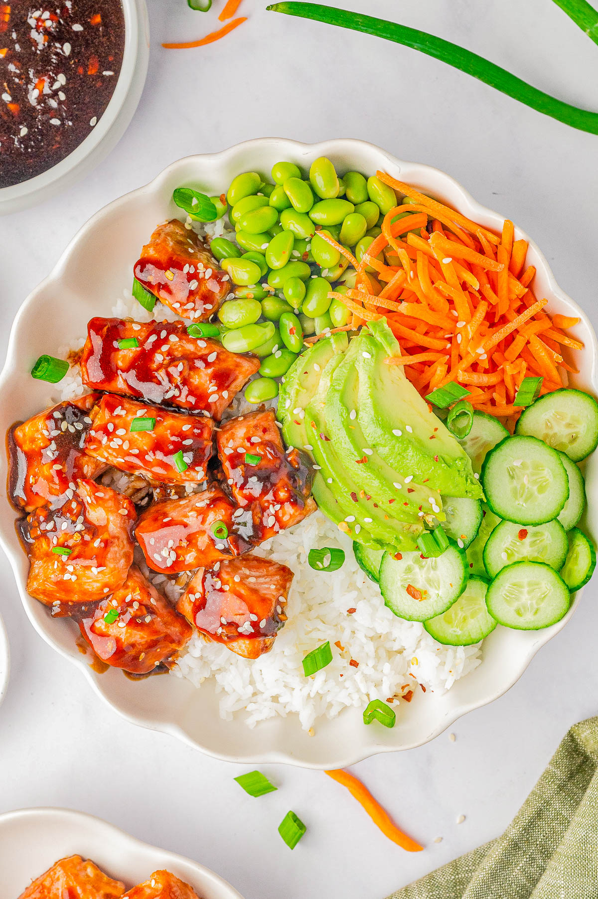 A bowl of rice topped with cubed glazed salmon, sliced avocado, cucumber, shredded carrots, and edamame, garnished with green onions and sesame seeds.