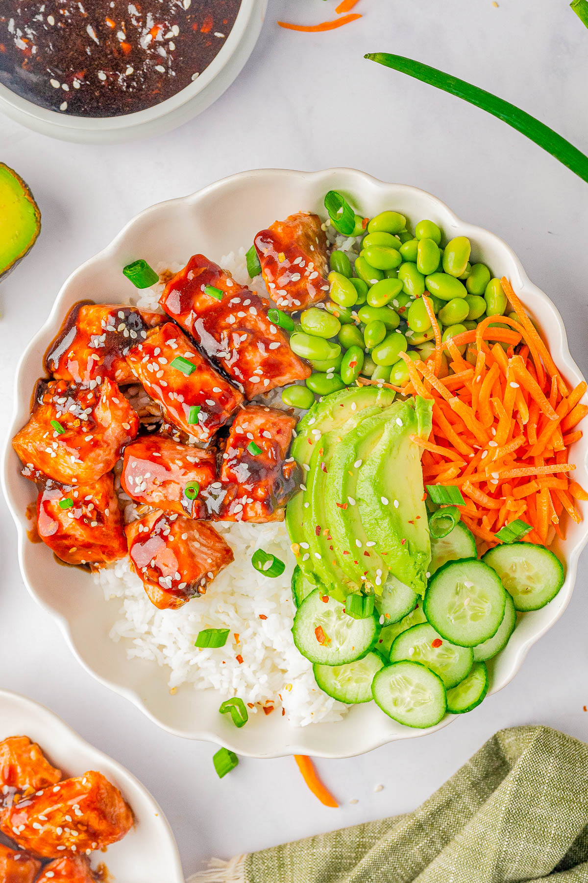 A bowl of white rice topped with cubed glazed protein, sliced avocado, cucumber, shredded carrots, edamame, and garnished with sesame seeds and green onions.