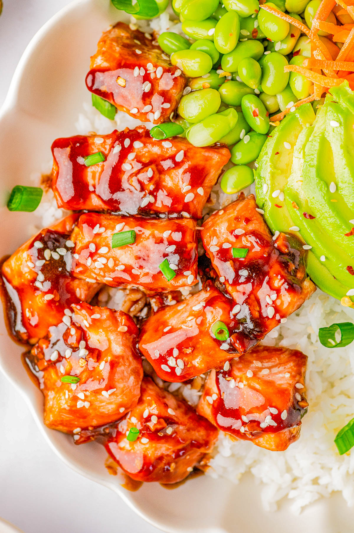 A bowl of rice topped with bite-sized pieces of glazed salmon sprinkled with sesame seeds, alongside edamame, sliced avocado, shredded carrots, and chopped green onions.