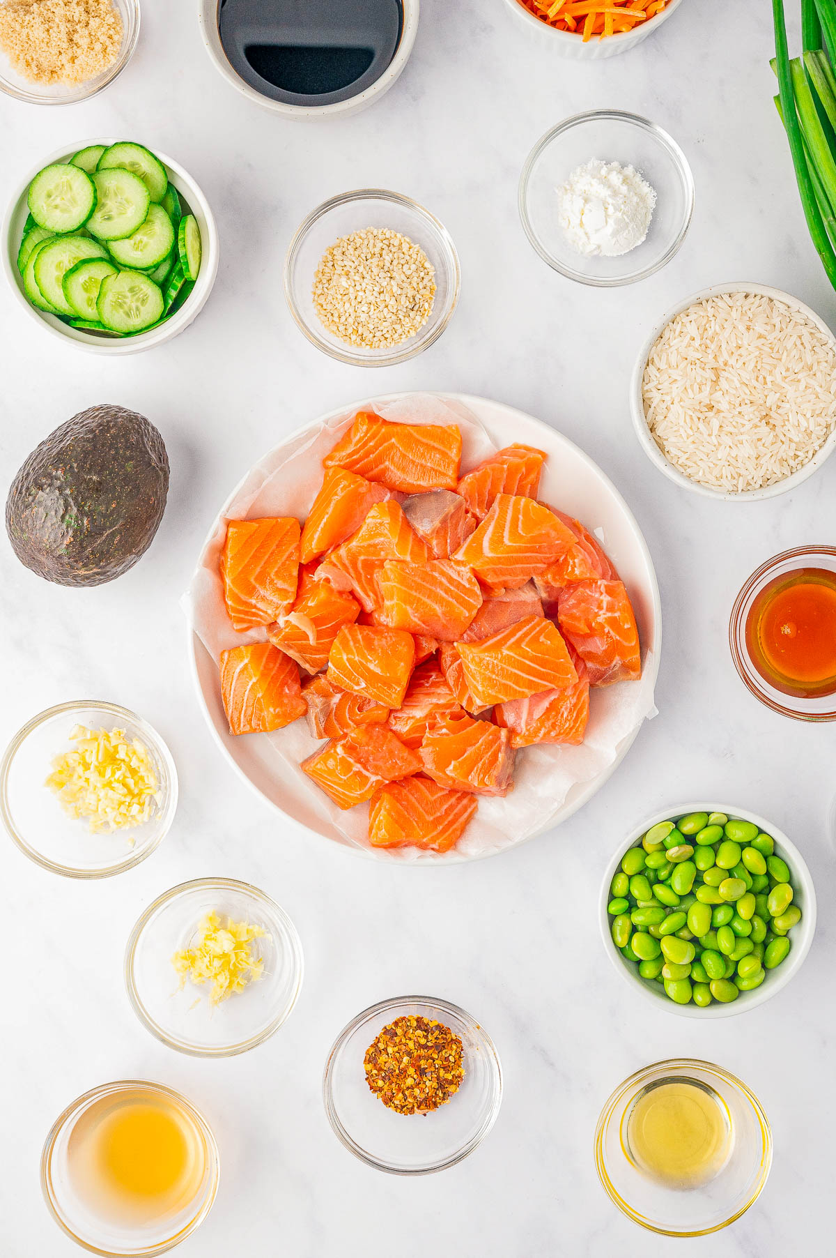 A plate of raw salmon pieces is surrounded by small bowls of various ingredients including sliced cucumbers, sesame seeds, rice, sliced avocado, grated ginger, edamame, sauce, and seasonings.