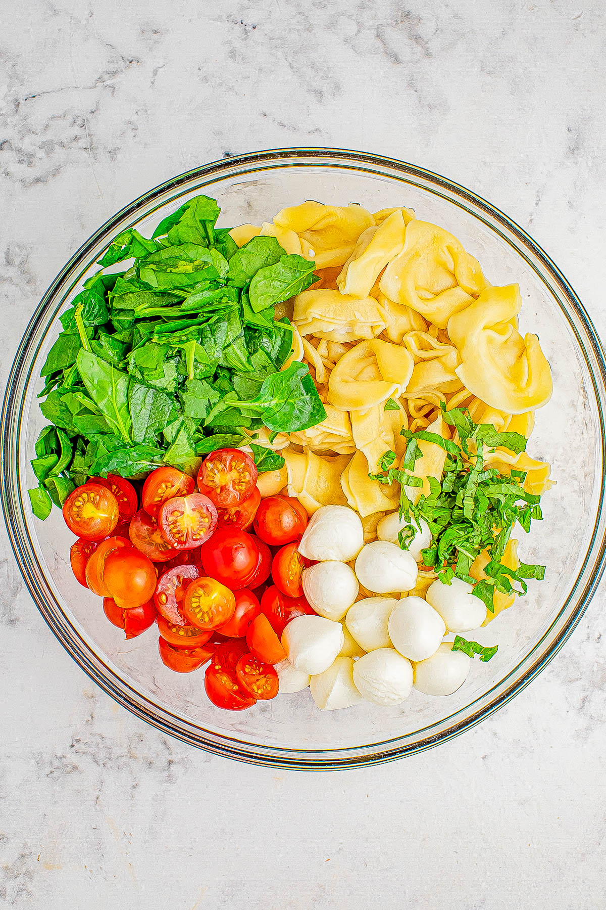 A glass bowl containing spinach, tortellini, sliced cherry tomatoes, mozzarella balls, and chopped basil arranged in sections on a marble countertop.