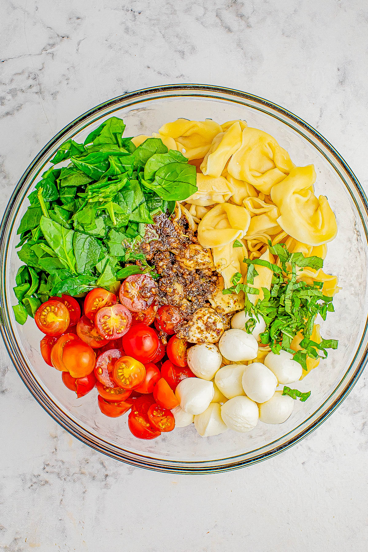 A clear bowl contains fresh spinach, tortellini, halved cherry tomatoes, mozzarella balls, and basil on a white marble surface.