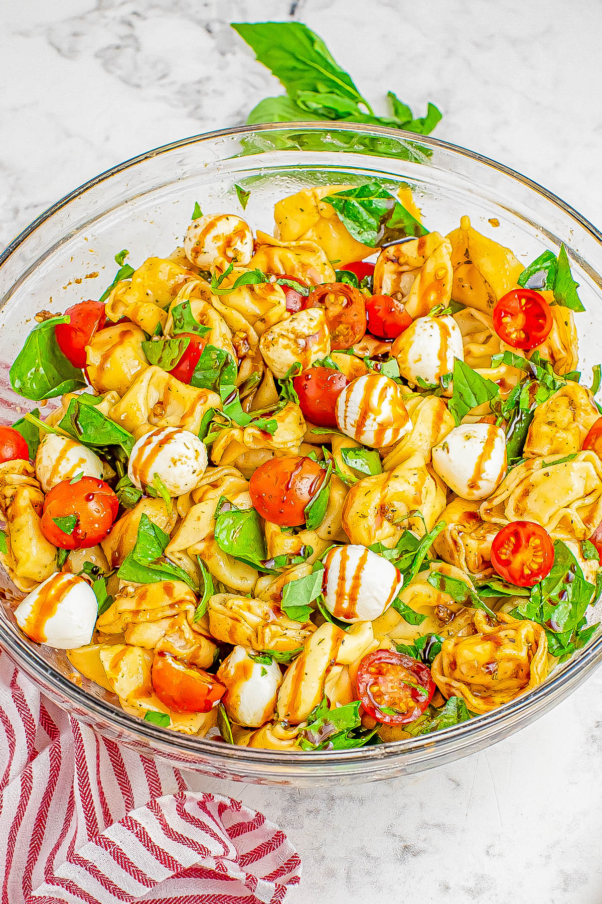 A bowl of tortellini pasta salad with cherry tomatoes, mozzarella balls, fresh basil, and a drizzle of balsamic glaze. The bowl sits on a white marble surface with a red-striped napkin nearby.