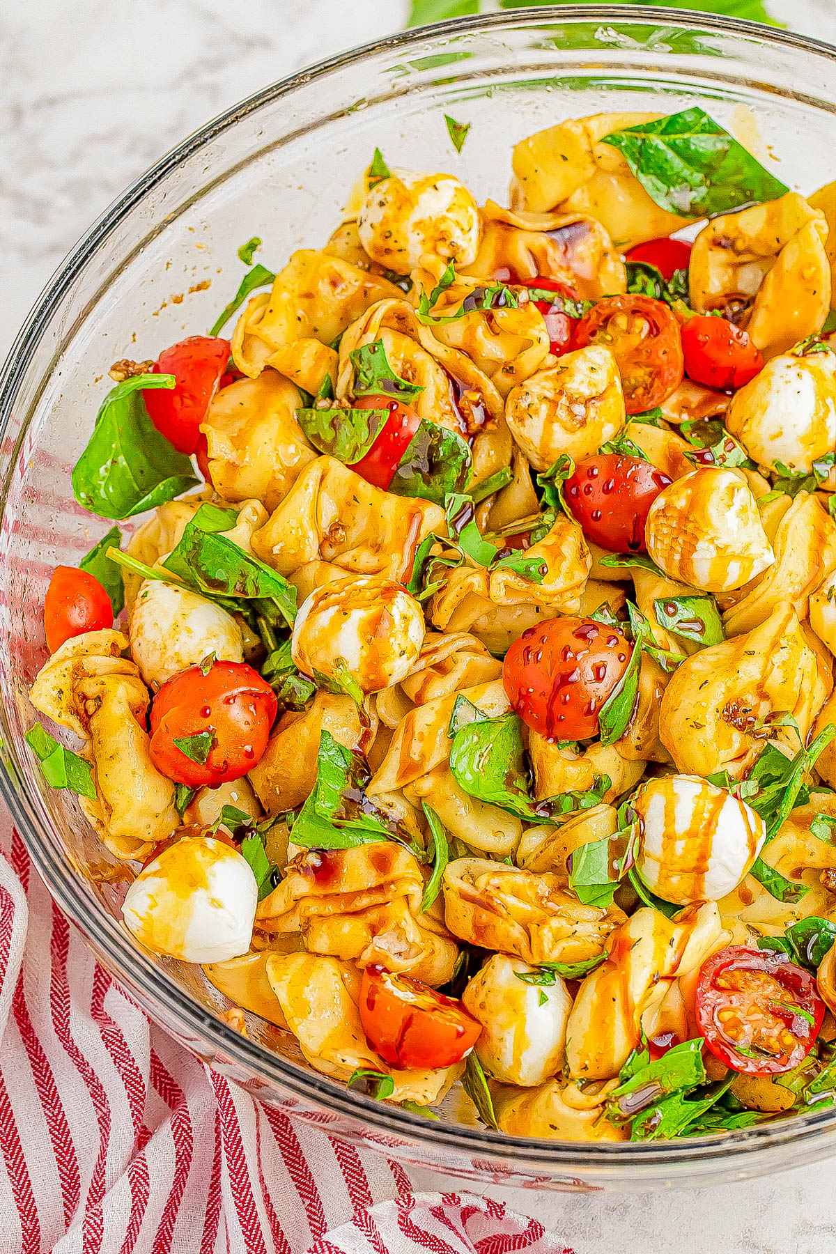 A glass bowl contains a vibrant tortellini pasta salad with tomatoes, mozzarella balls, and fresh basil, drizzled with balsamic glaze. A red and white striped cloth is partially visible underneath.