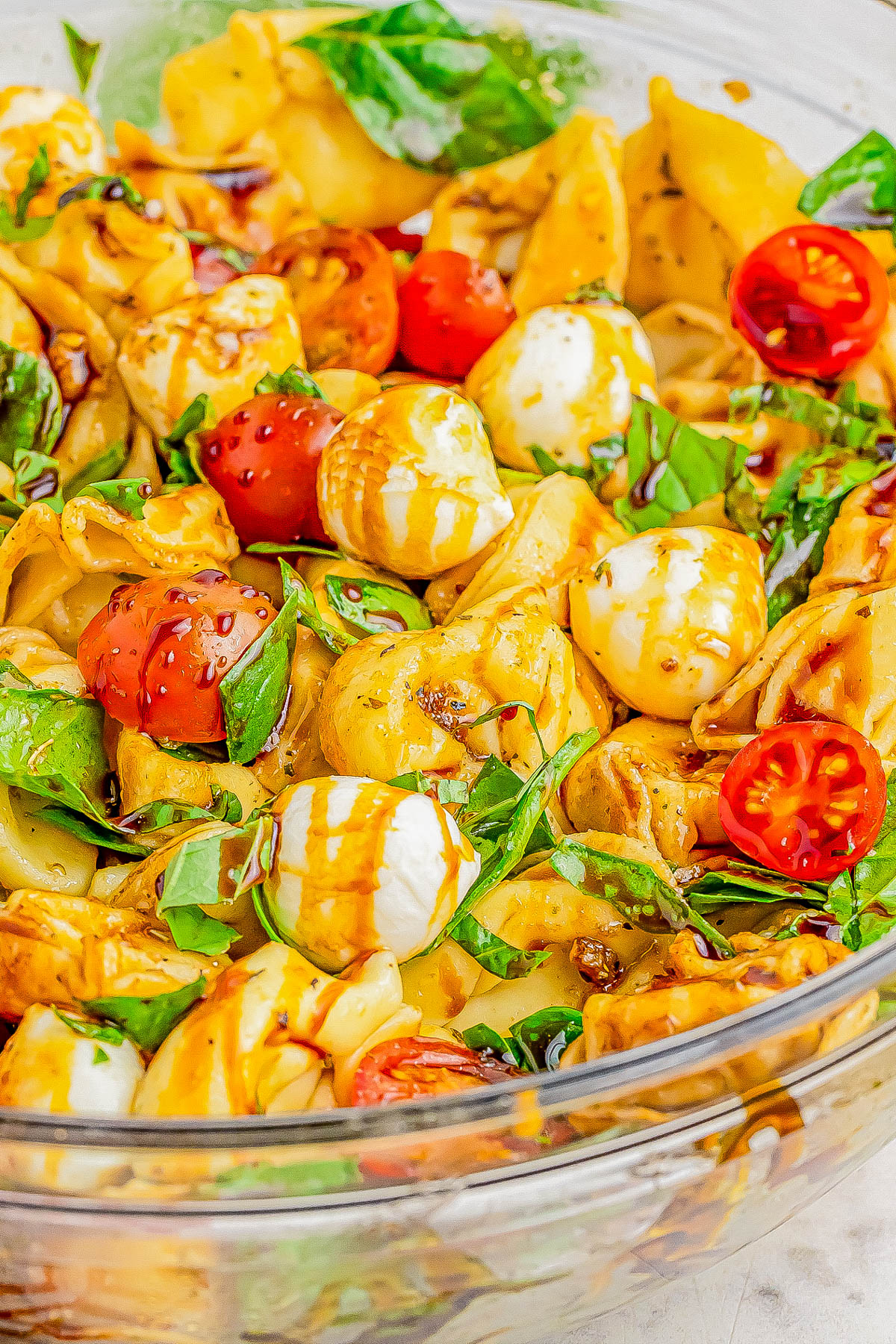 A glass bowl filled with tortellini pasta, cherry tomatoes, mozzarella balls, fresh basil leaves, and drizzled with balsamic glaze.