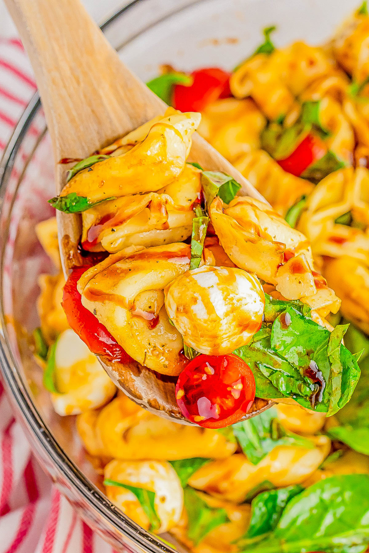 A wooden spoon holding a portion of tortellini pasta salad with tomatoes, mozzarella, spinach, and basil.