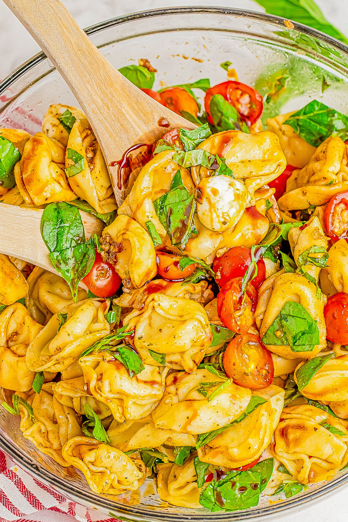 A close-up of a bowl of tortellini pasta salad mixed with cherry tomatoes, fresh spinach, and mozzarella balls, being stirred with two wooden spoons.