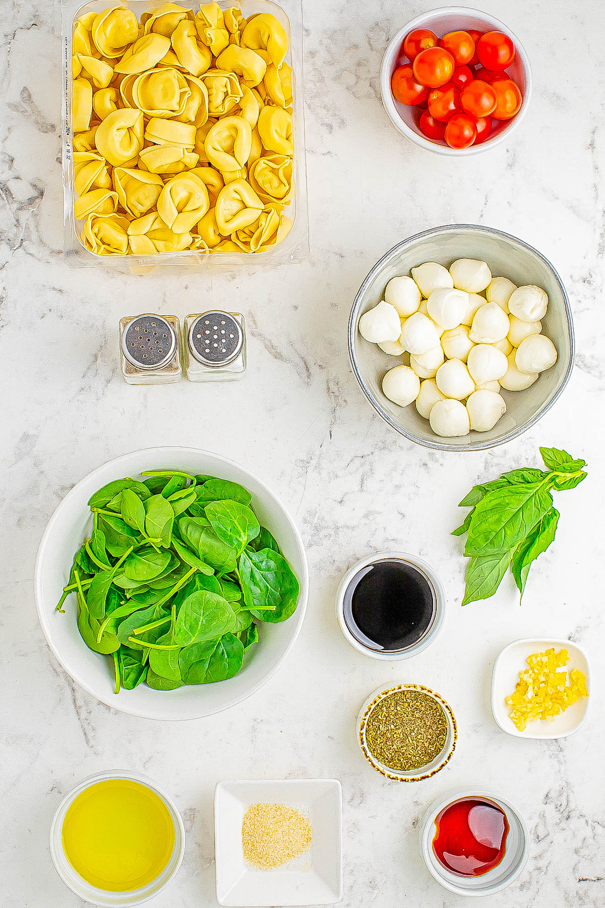 Top-down view of pasta salad ingredients including tortellini, cherry tomatoes, mozzarella balls, spinach, olive oil, balsamic vinegar, garlic, Italian seasoning, and fresh basil on a marble surface.