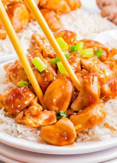 A plate of white rice topped with chunks of saucy chicken garnished with sesame seeds and sliced green onions, being picked up with chopsticks.