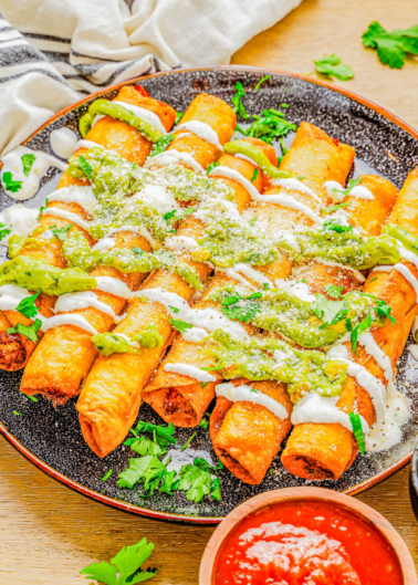 A plate of crispy taquitos topped with green sauce, cream, crumbled cheese, and garnished with fresh cilantro. A small bowl of red sauce is in the foreground.