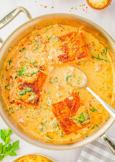 A pan filled with creamy sauce, pieces of cooked salmon, spinach, and herbs. A spoon rests inside the sauce, and some parsley and a dish of grated cheese are nearby.