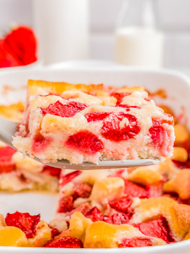 Close-up of a spatula holding a slice of strawberry cobbler, with the rest of the dessert visible in the background. The cobbler has a golden crust interspersed with pieces of fresh strawberries.