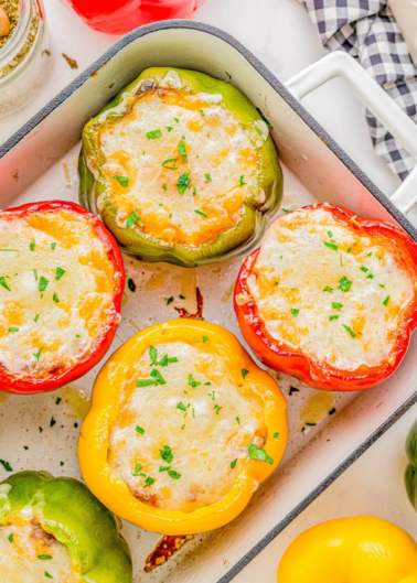 Four stuffed bell peppers with melted cheese, in green, red, and yellow, baked and garnished with herbs, in a white baking dish.