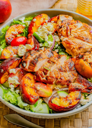 A bowl of salad featuring grilled chicken breast, mixed greens, sliced peaches, cucumbers, and crumbled blue cheese, set on a wooden placemat with a fork nearby.