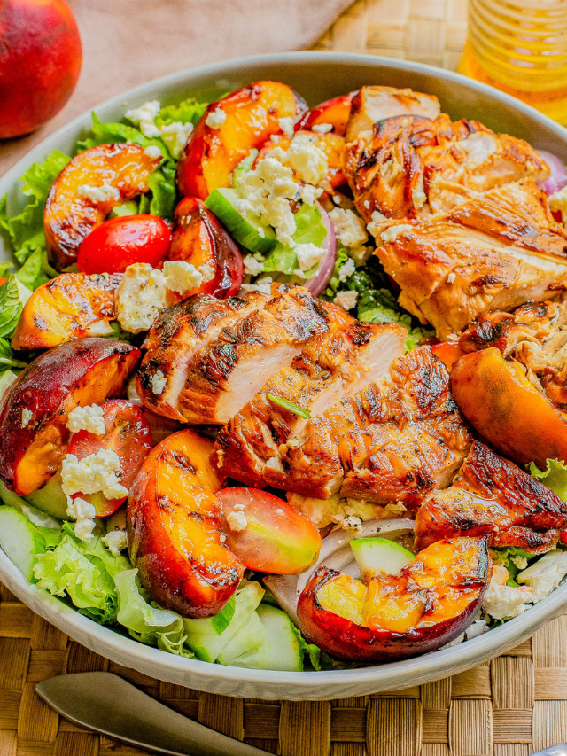 A bowl of salad featuring grilled chicken breast, mixed greens, sliced peaches, cucumbers, and crumbled blue cheese, set on a wooden placemat with a fork nearby.