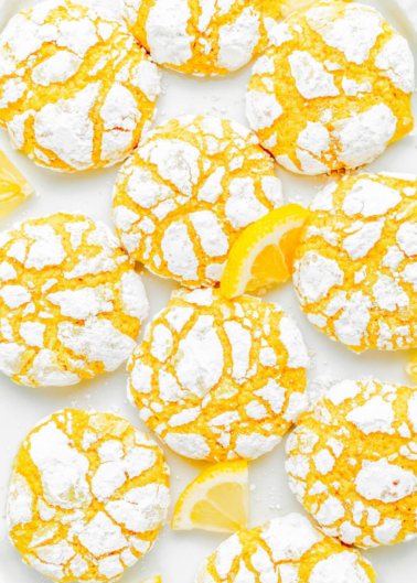 A close-up of lemon crinkle cookies dusted with powdered sugar, arranged in a pattern on a white surface, alongside lemon slices.