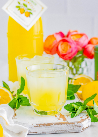 A glass of lemonade with ice cubes on a white wooden surface, accompanied by lemon slices and mint leaves. A bottle of lemonade and a vase with pink and orange flowers are in the background.