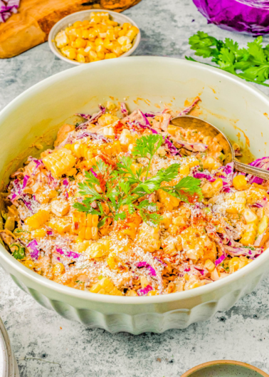 A large bowl filled with a mixed salad containing corn, grated cheese, cabbage, and vegetables, garnished with a sprig of parsley, and two serving spoons. Small bowls of corn and parsley in the background.