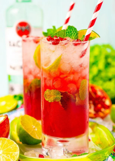 Two tall glasses of a red cocktail garnished with lime slices, mint leaves, and red straws. A bottle, lime, and pomegranate are in the background.