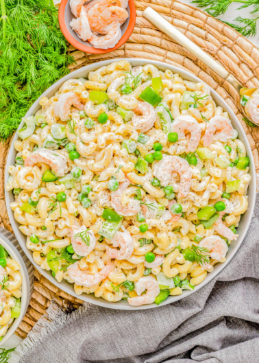 A bowl of macaroni salad with shrimp, peas, bell pepper, and dill, placed on a woven mat beside fresh dill and a small bowl of shrimp. A gray cloth and fork are also visible.