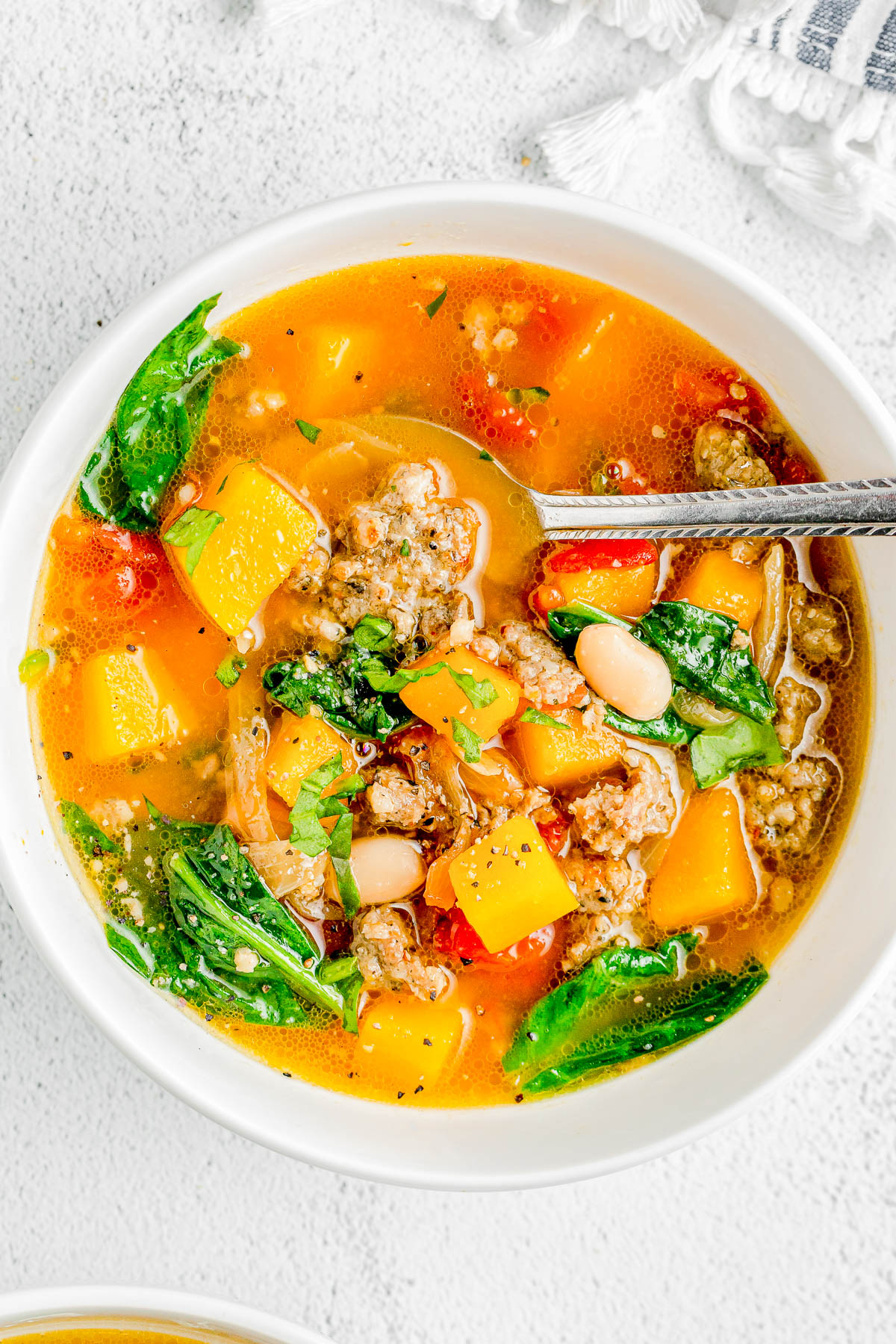 A bowl of soup containing ground meat, diced butternut squash, white beans, spinach, and tomatoes with a spoon inside.