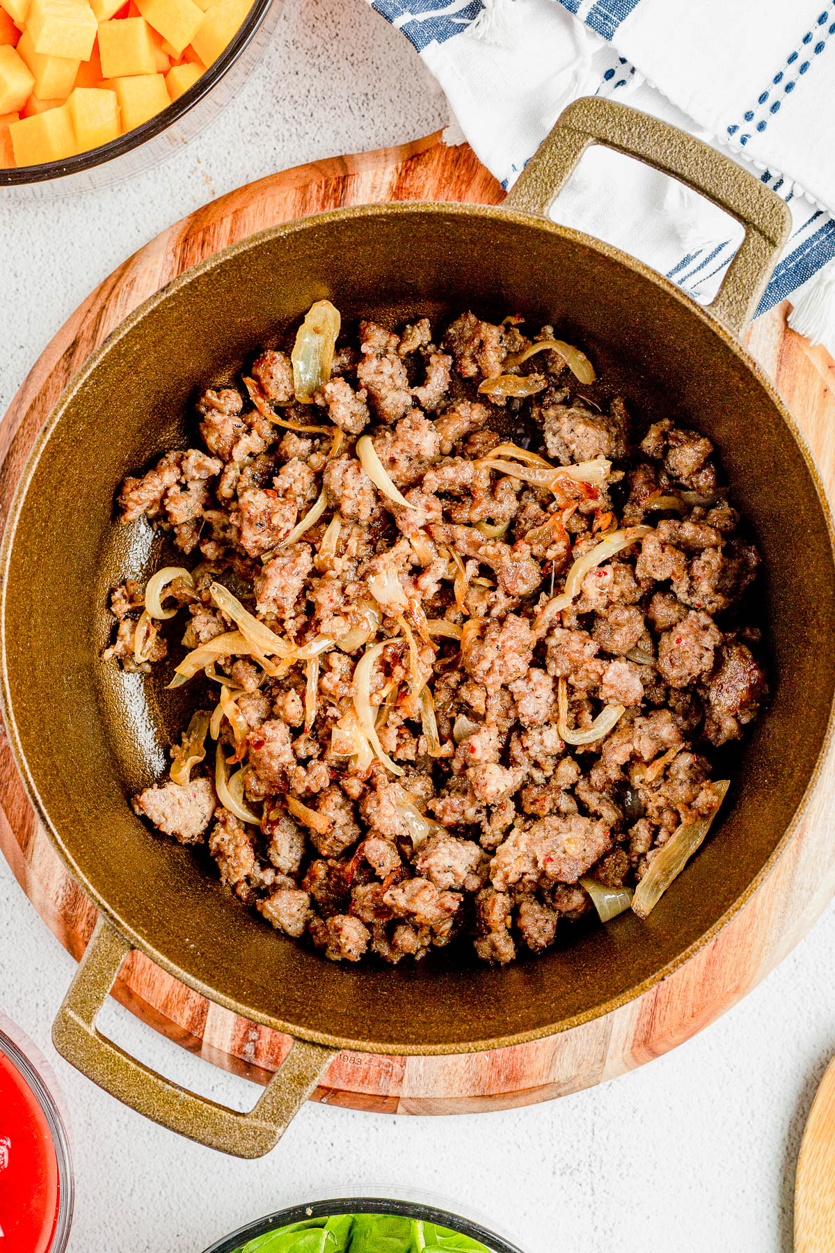 A pot of cooked ground sausage mixed with sautéed onions is placed on a wooden board, surrounded by bowls of diced orange vegetables, spinach leaves, and a red liquid.