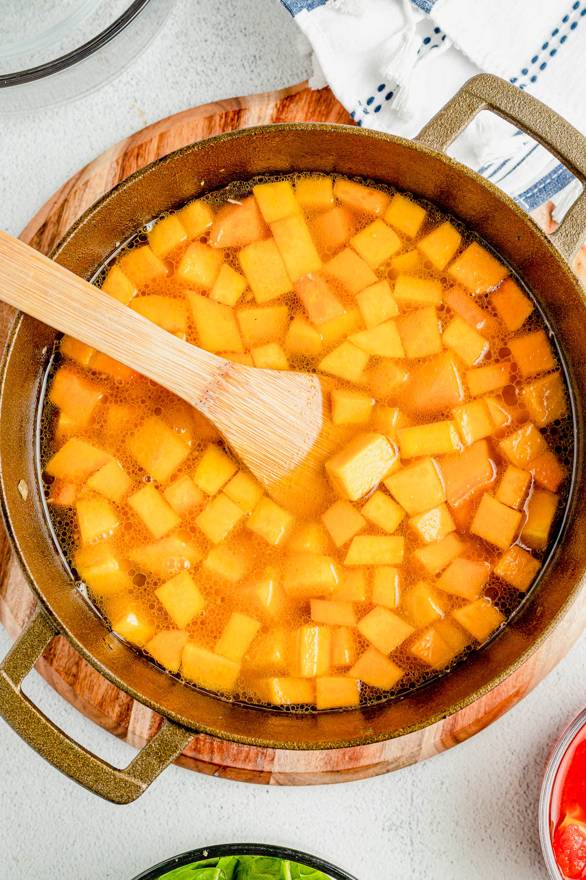 A pot of diced squash is being stirred with a wooden spoon. The pot is placed on a wooden board, and there are other ingredients nearby.