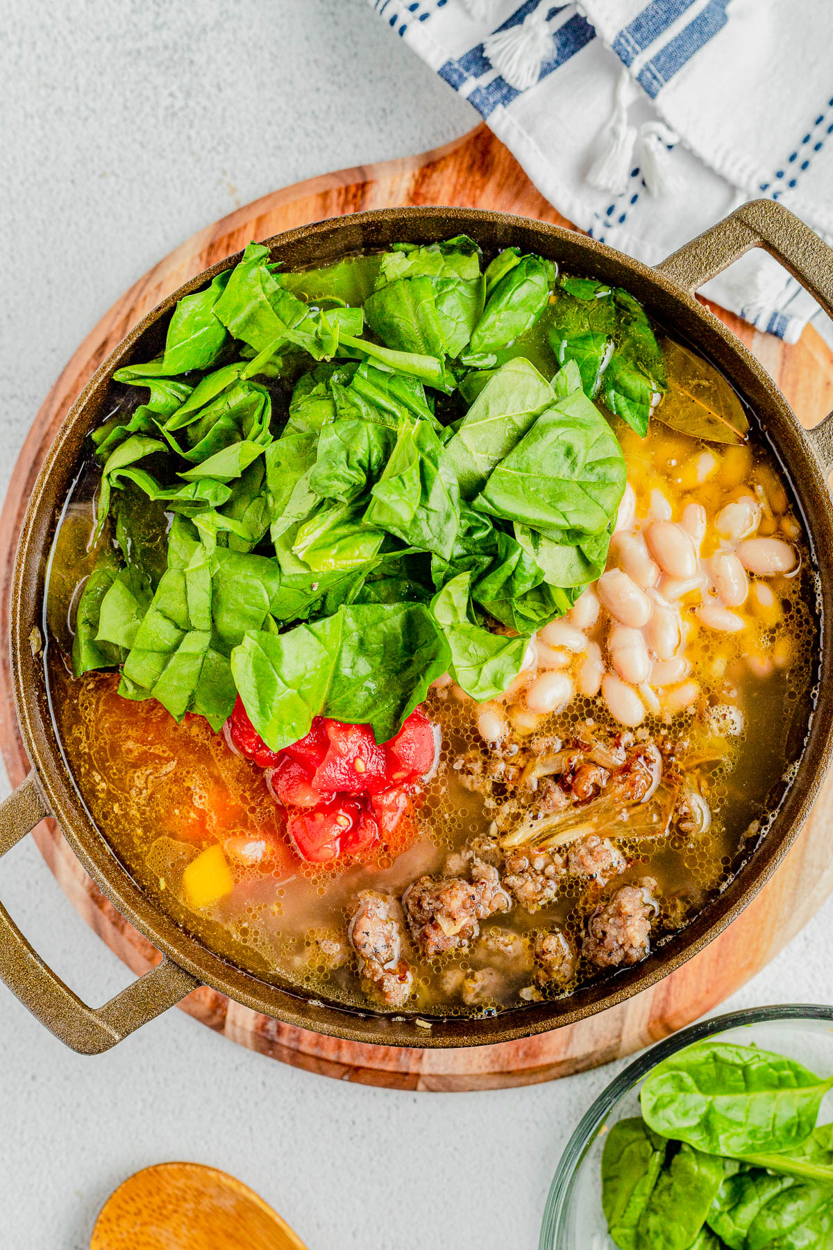 A pot filled with soup containing white beans, ground meat, chopped tomatoes, and fresh spinach, placed on a wooden board with a small bowl of spinach and a wooden spoon nearby.