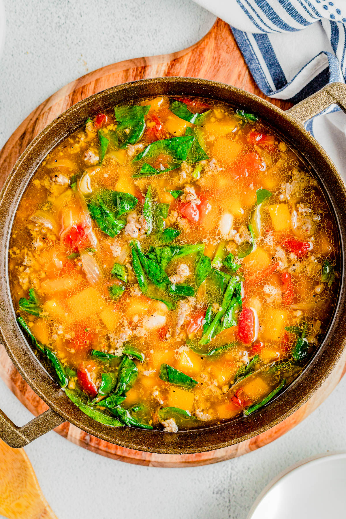 A pot of vegetable soup filled with a variety of colorful ingredients, including greens, tomatoes, and squash, sits on a wooden board next to a white plate and blue-striped cloth.