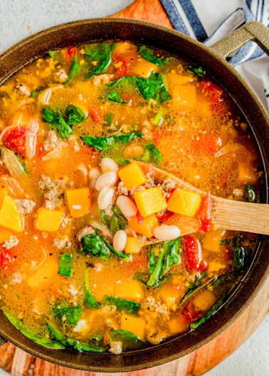 A pot of soup containing chunks of squash, white beans, spinach, tomatoes, and ground meat, with a wooden spoon scooping some out.