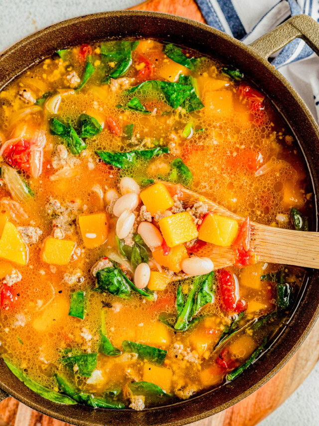 A pot of soup containing chunks of squash, white beans, spinach, tomatoes, and ground meat, with a wooden spoon scooping some out.