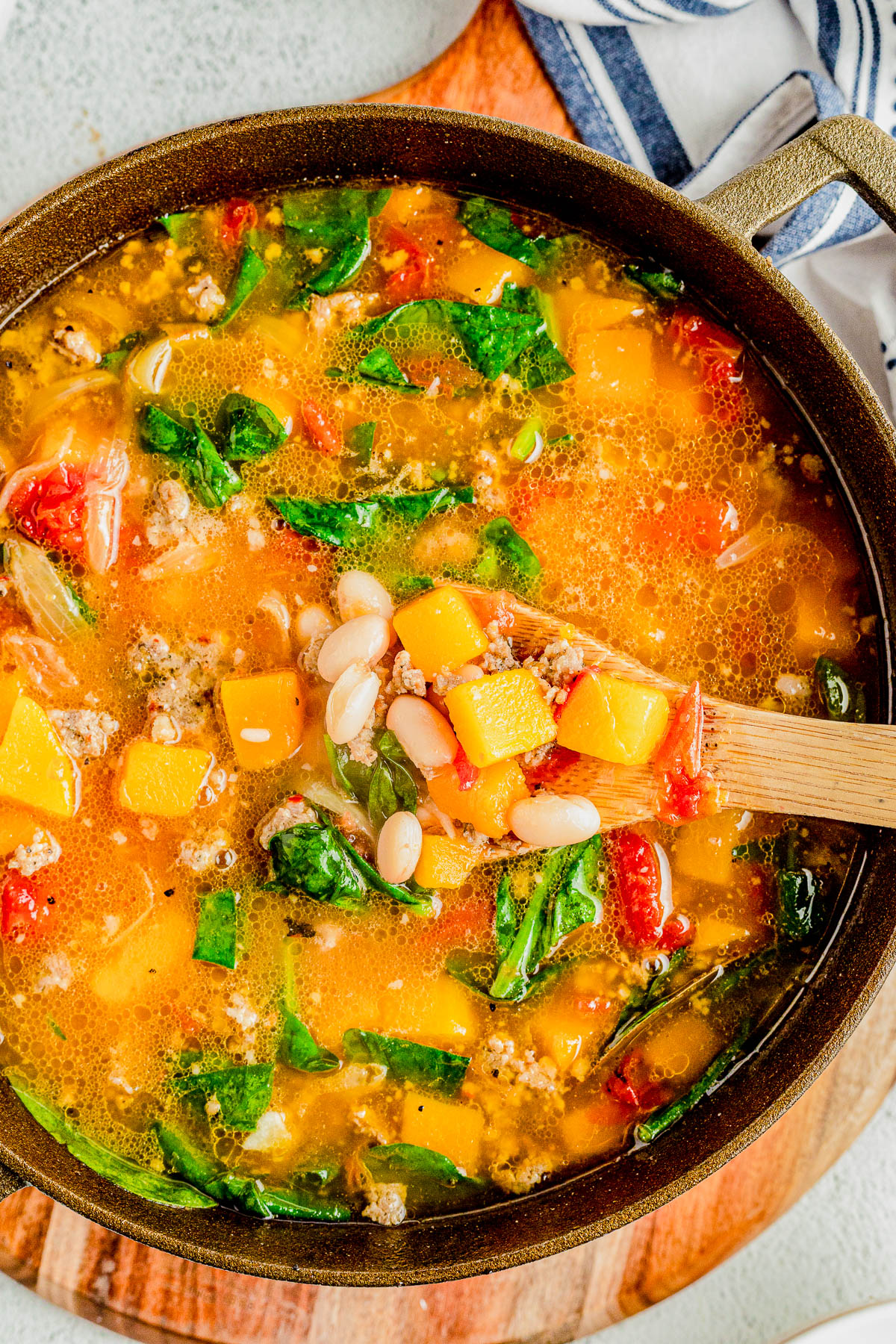 A pot of soup containing chunks of squash, white beans, spinach, tomatoes, and ground meat, with a wooden spoon scooping some out.