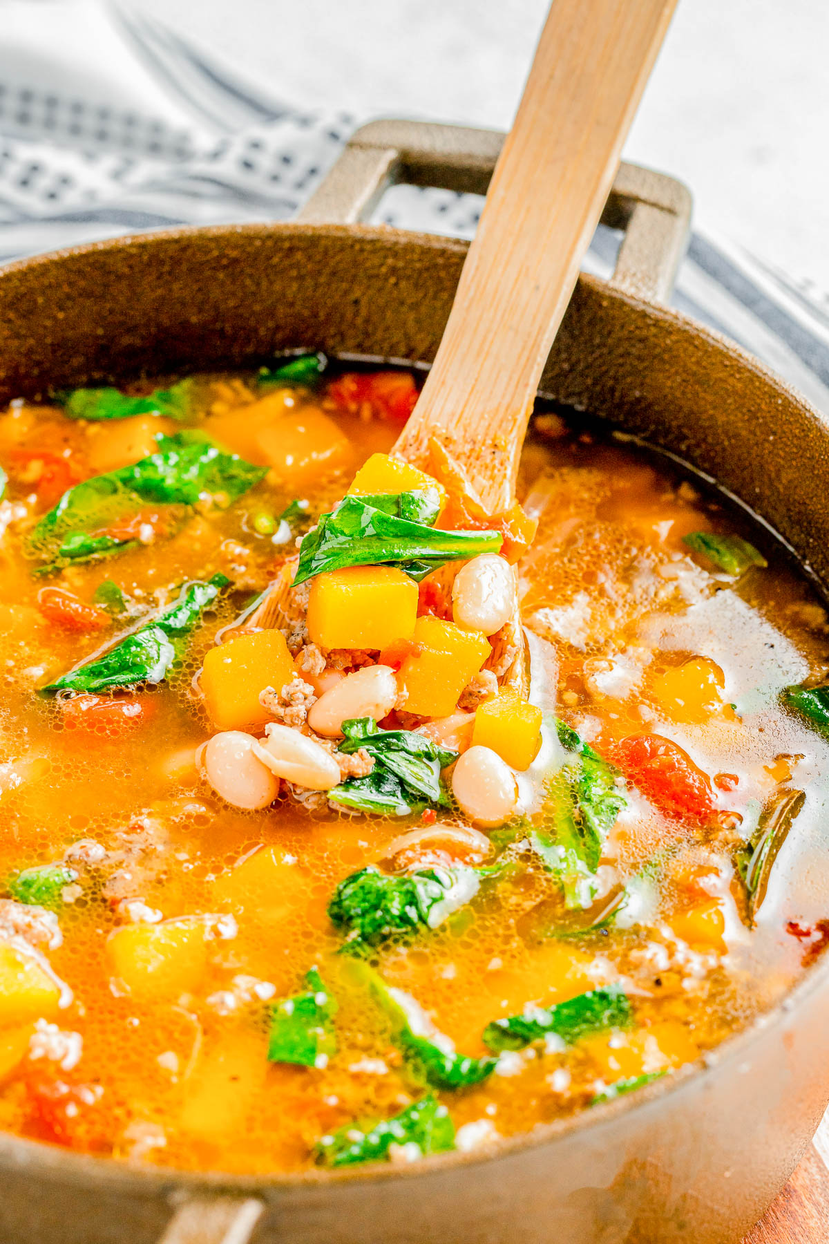 A pot of vegetable soup with white beans, diced yellow squash, spinach, and tomatoes, with a wooden spoon stirring the contents.