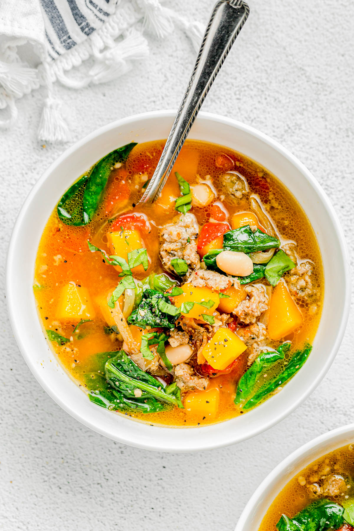 A bowl of colorful soup with chunks of meat, yellow squash, leafy greens, beans, and herbs, with a spoon resting in it.