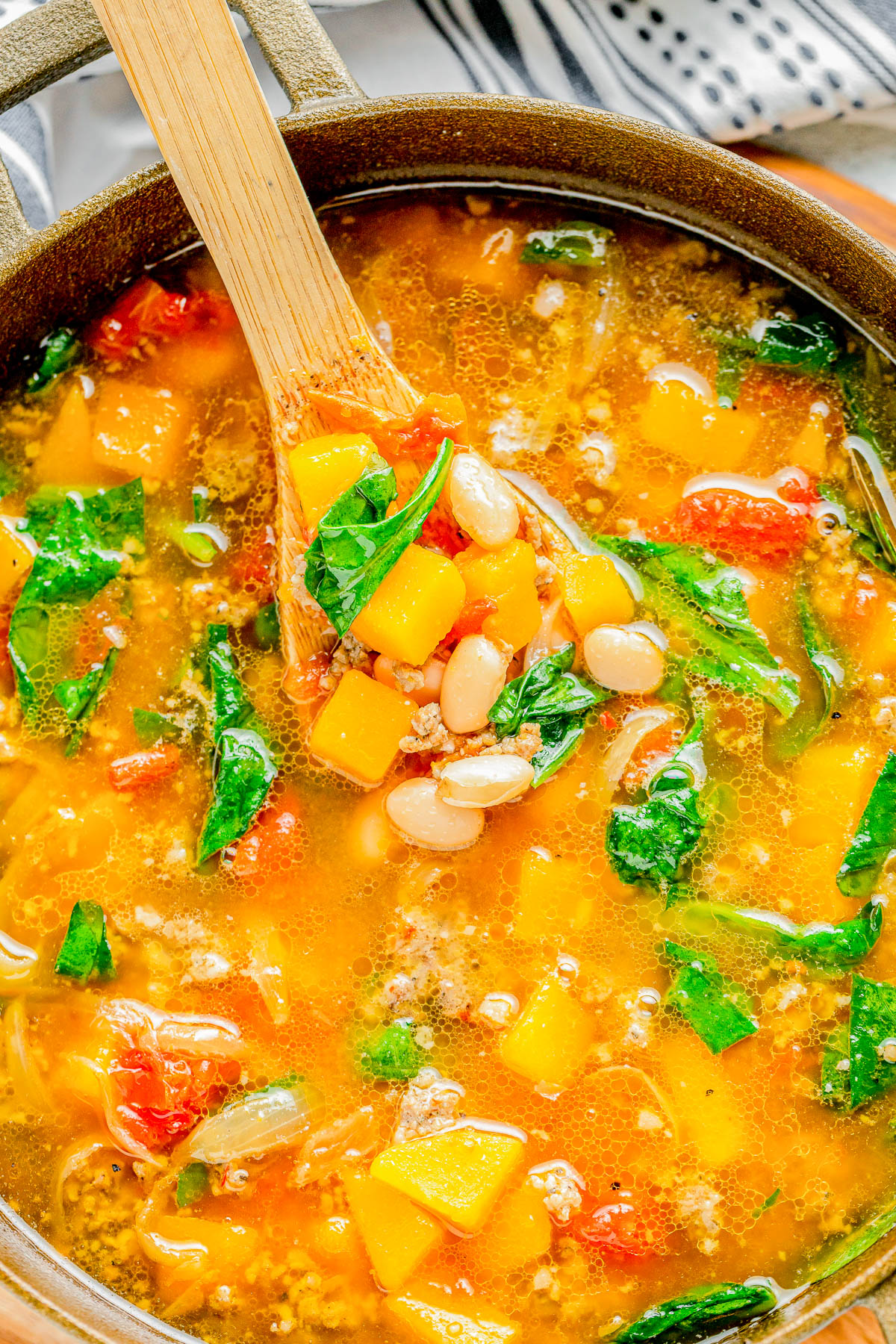 A pot of vegetable soup being stirred with a wooden spoon. The soup contains chunks of squash, white beans, spinach, tomatoes, and broth.