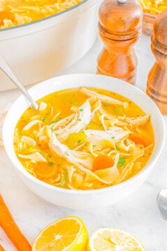 A bowl of chicken noodle soup with shredded chicken, noodles, and carrots, garnished with herbs, sits on a white surface next to a pot, two wooden pepper grinders, and halved lemons.